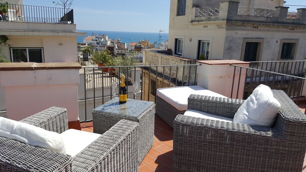 A balcony with wicker furniture, a bottle of champagne, and a glass. The balcony overlooks a cityscape with buildings and a view of the sea. - The Sky penthouse in the center of town with panoramic views and sunny terrace (Sitges)