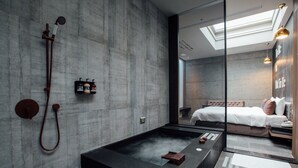 A modern bathroom with a large black bathtub, a shower, and a glass-enclosed bedroom with a bed and a nightstand.