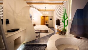 A modern hotel room with a bed, a small seating area, a cactus decoration, and a unique ceiling design.
