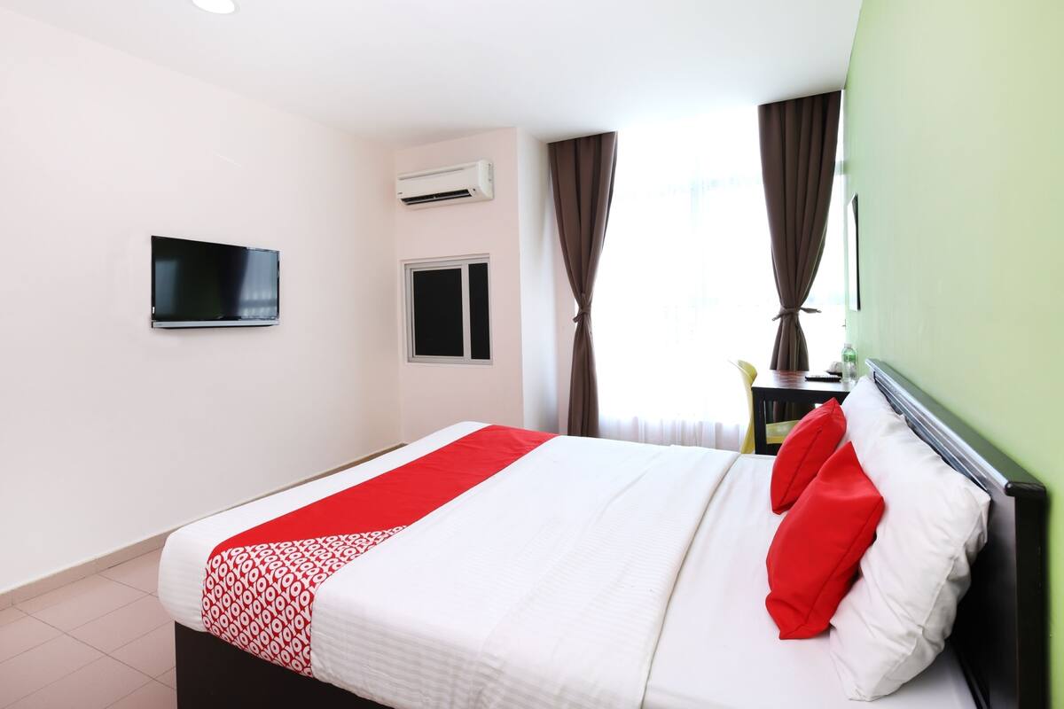 A hotel room with a bed, a television mounted on the wall, an air conditioning unit, a window with curtains, and a small table with a bottle.