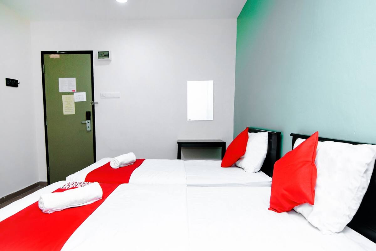 A hotel room with two beds, red and white bedding, a small bedside table, a green door, and a white wall with a window.