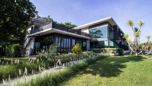 Een modern herenhuis met twee verdiepingen en een balkon, omgeven door weelderige begroeiing en een verzorgd gazon.