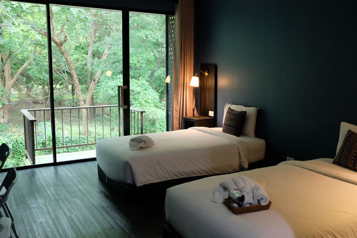 A hotel room with two beds, a balcony with a view of trees, a desk with a chair, and a nightstand with a lamp.