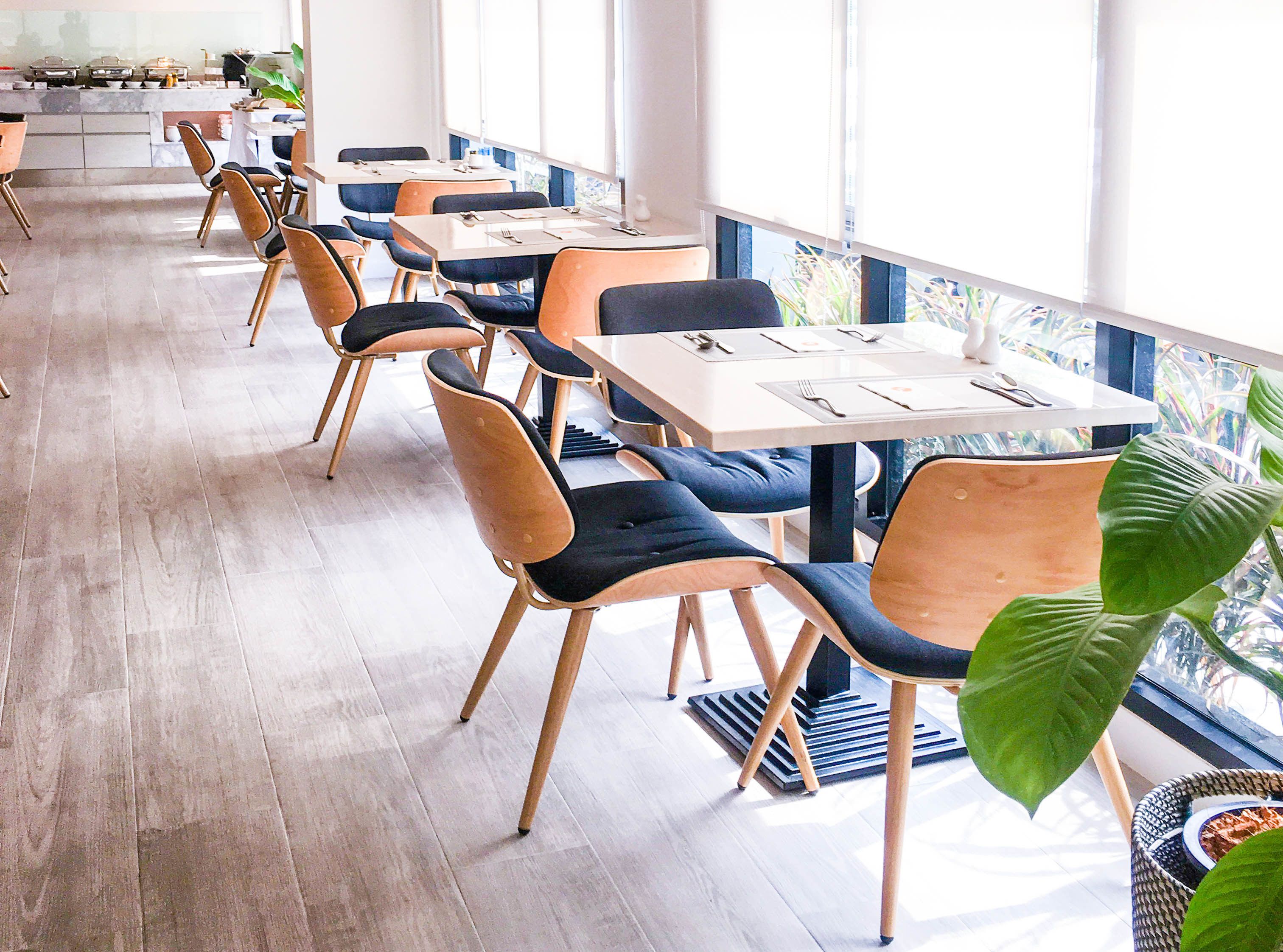 A modern dining area with wooden tables and chairs, large windows, and potted plants.