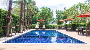 A swimming pool with lounge chairs and red umbrellas, surrounded by palm trees and greenery. - Fortune Hotel Buriram (Buriram)