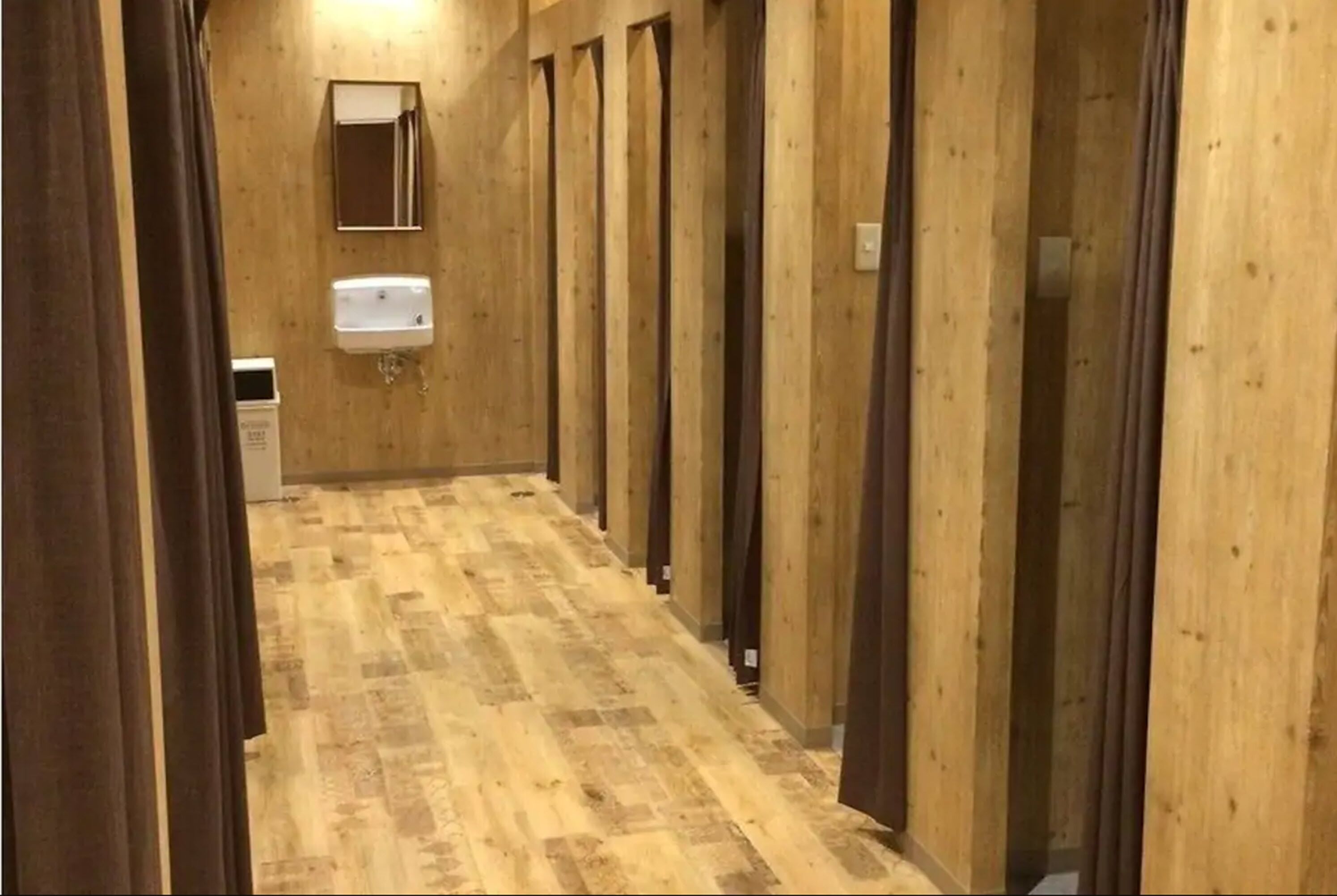 Un couloir aux murs en bois et au sol carrelé, qui mène à des boxes individuels équipés de lavabos.