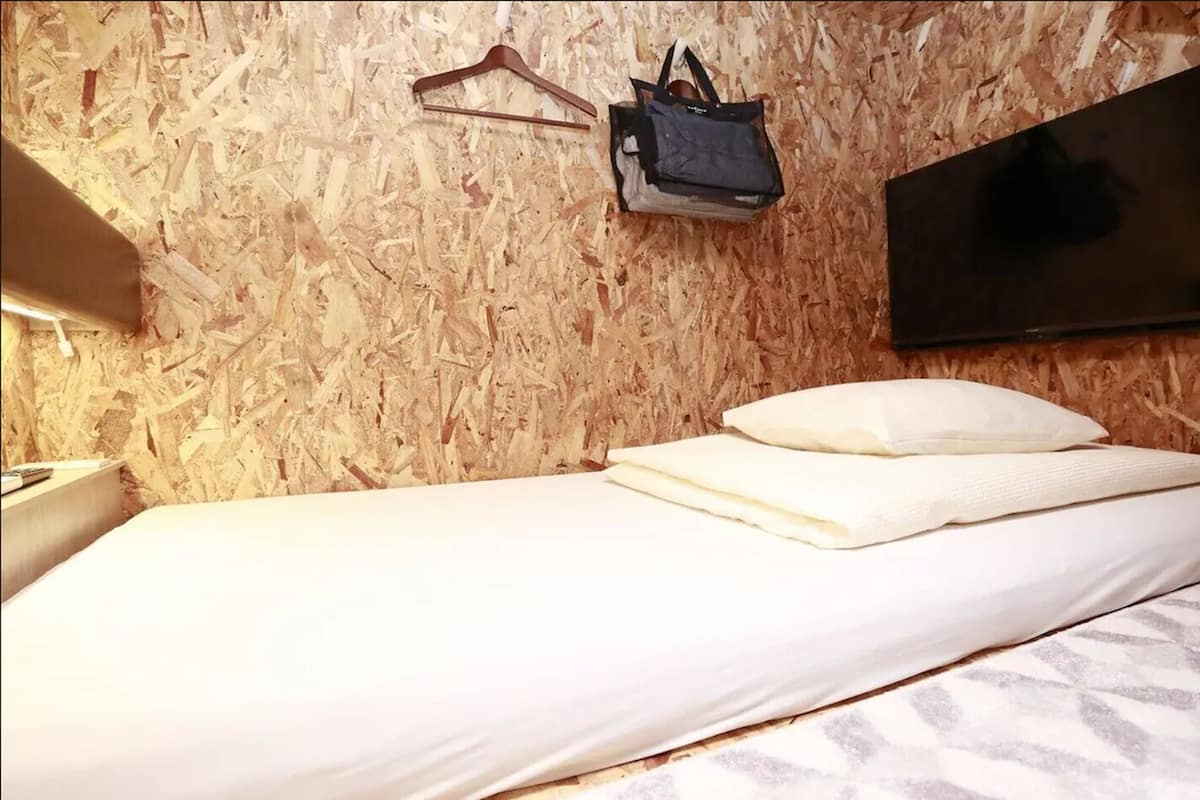 A room with a bed, a TV mounted on the wall, a hanging bag, and a wooden wall.