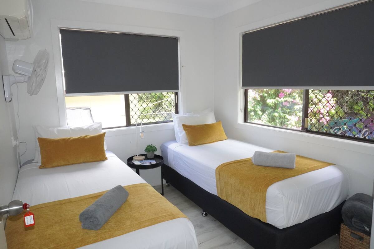 Une chambre d’hôtel avec deux lits, chacun doté d’un matelas blanc et d’un plaid jaune. Une petite table avec une lampe et une plante se trouve entre les deux lits.