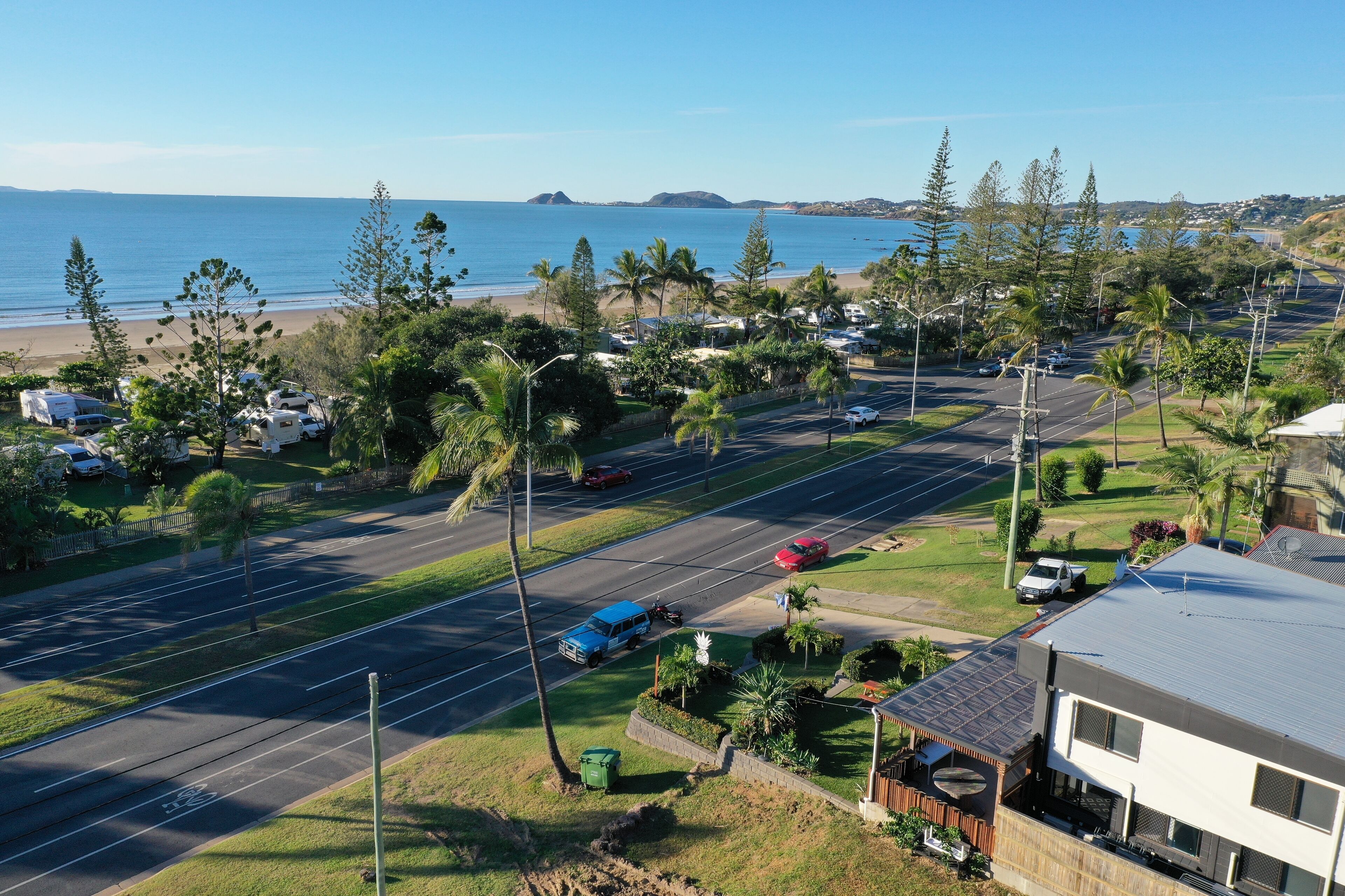 A coastal road with a beach, parked cars, and a modern house with a balcony.