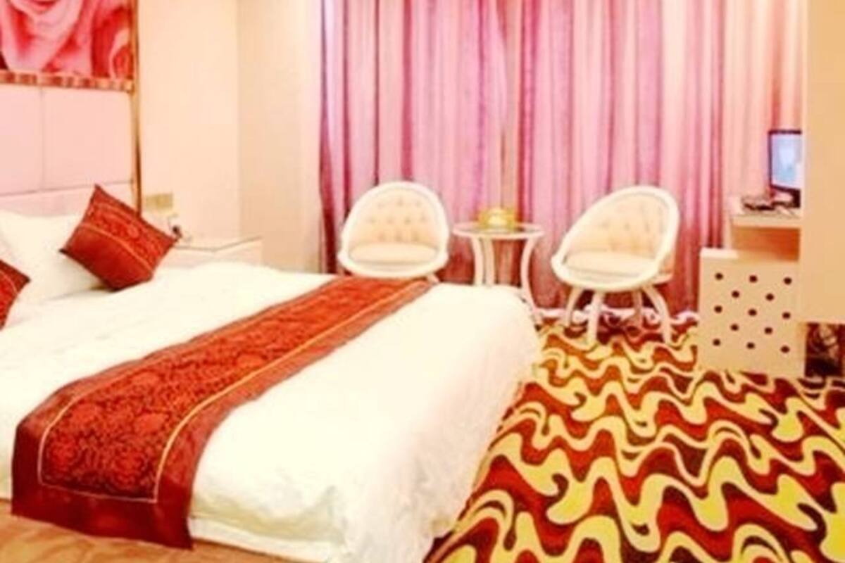 Quarto de hotel com uma cama, um tapete com padrão vermelho e amarelo, uma cortina rosa e duas cadeiras brancas.