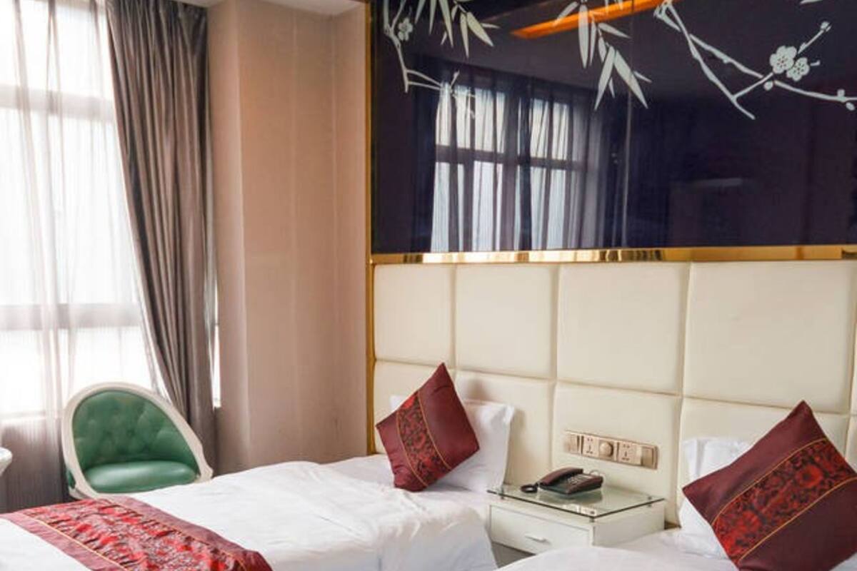 Quarto de hotel com duas camas, uma cadeira verde, uma mesa de cabeceira e uma janela grande com cortinas.