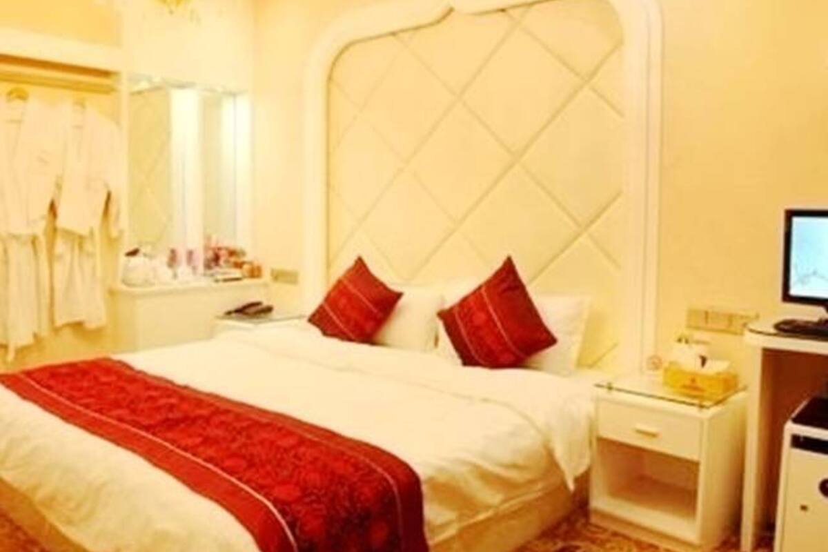 Quarto de hotel com uma cama grande, um candelabro, uma mesinha de cabeceira e uma escrivaninha com um computador.