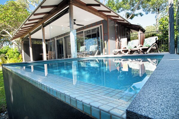 Una piscina con un patio cubierto, sillas de descanso y una habitación con paredes de vidrio y un ventilador de techo.