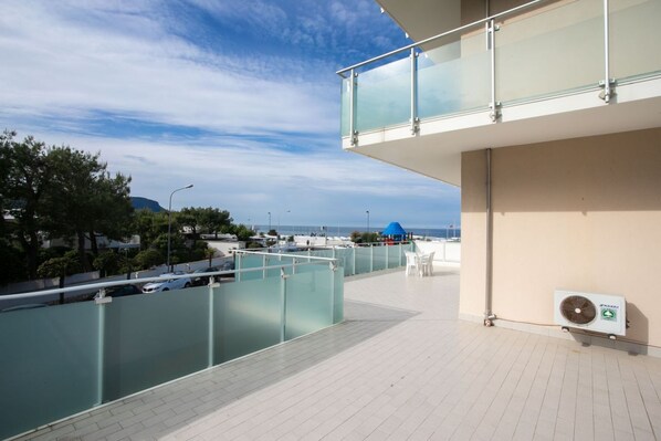 A rooftop terrace with a glass balustrade, a fan, and a view of the sea and cityscape.