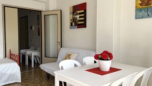 Una habitació d'hotel amb un llit, un sofà blanc, una taula de menjador amb una protecció vermella i un vas amb flors vermelles.