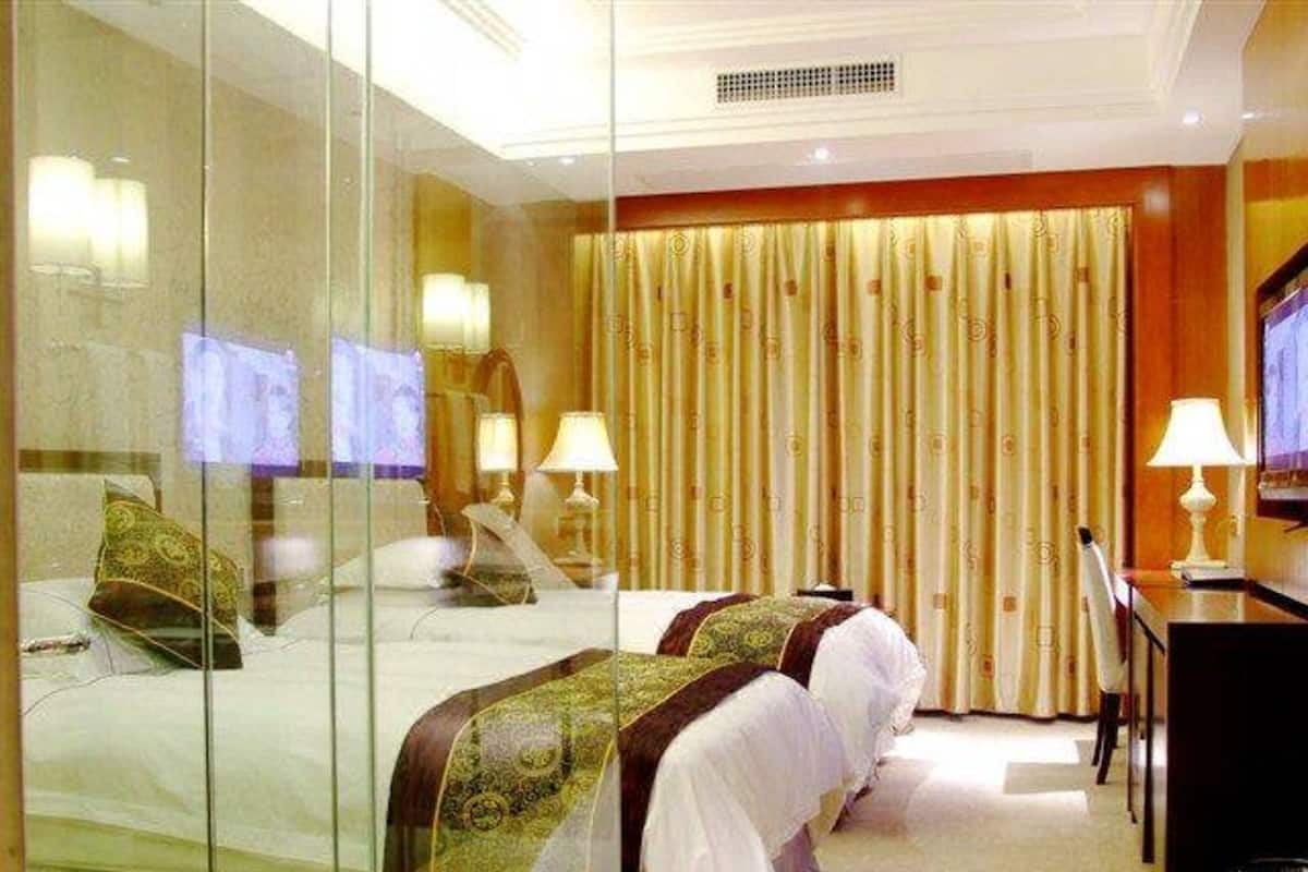 Une chambre d’hôtel avec une cloison en verre, deux lits avec des couvre-lits à motifs, un bureau avec une lampe et un grand rideau.