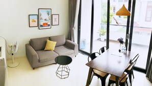 Un salon moderne avec un canapé gris, une table basse ronde et une table à manger avec des chaises. Des tableaux encadrés ornent le mur, un ventilateur sur pied est présent, et de grandes fenêtres offrent une vue sur l’extérieur.