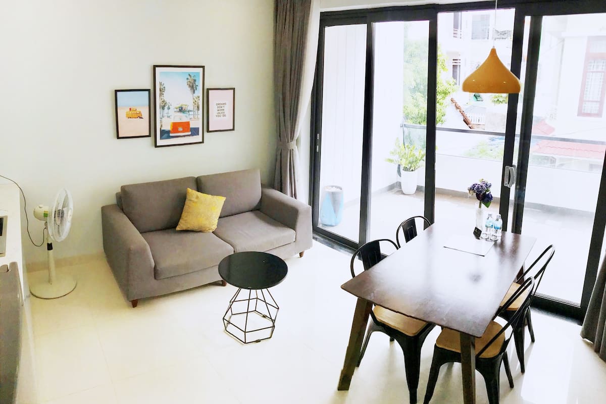 Un salon moderne avec un canapé gris, une table basse ronde et une table à manger avec des chaises. Des tableaux encadrés ornent le mur, un ventilateur sur pied est présent, et de grandes fenêtres offrent une vue sur l’extérieur.