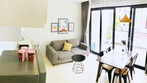 Un salon moderne avec un canapé gris, une table à manger avec des chaises et une table basse. Des tableaux encadrés ornent le mur, et une fenêtre est agrémentée de rideaux.