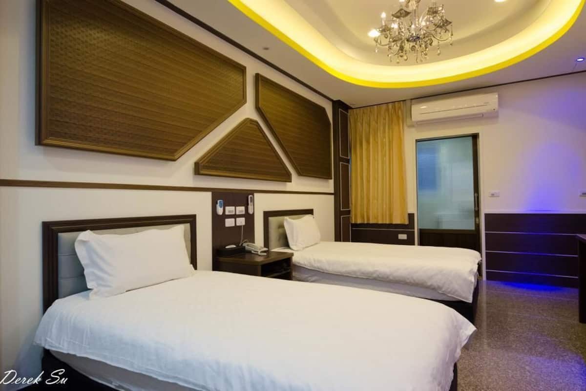 A hotel room with two beds, a chandelier, an air conditioner, and a window with curtains.