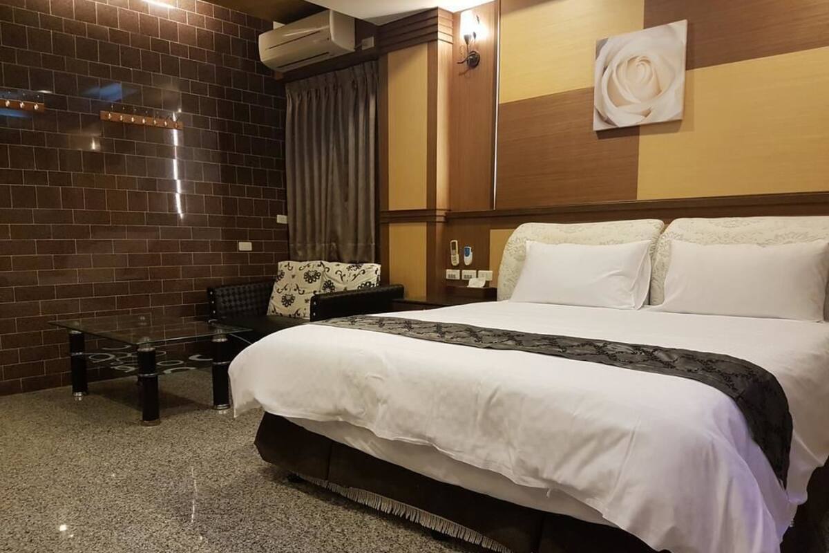 A hotel room with a large bed, a small table, a bench, and a wall-mounted air conditioner.