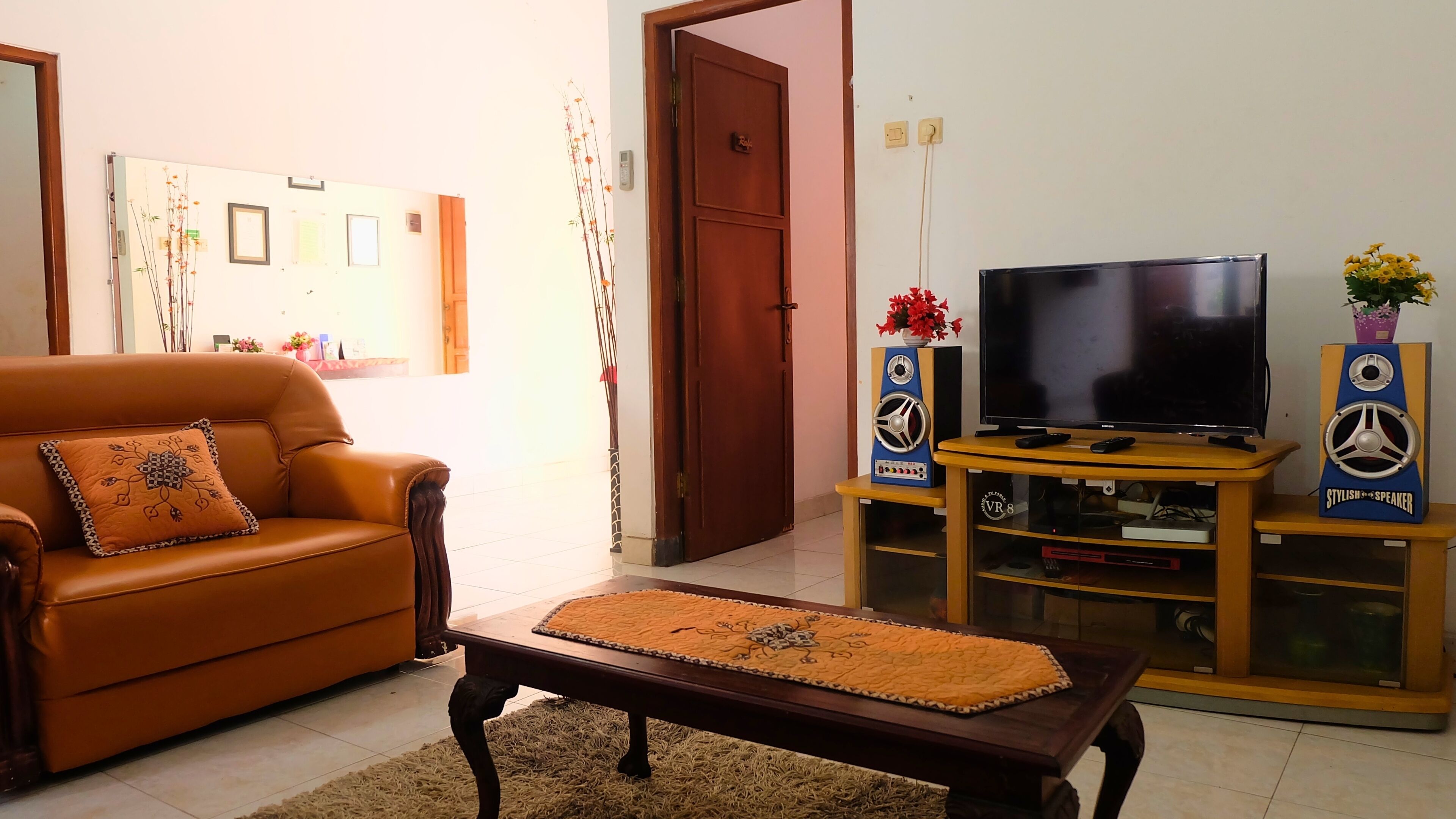 A living room with a brown leather sofa, a wooden coffee table, a flat-screen TV on a wooden stand, and two speakers.