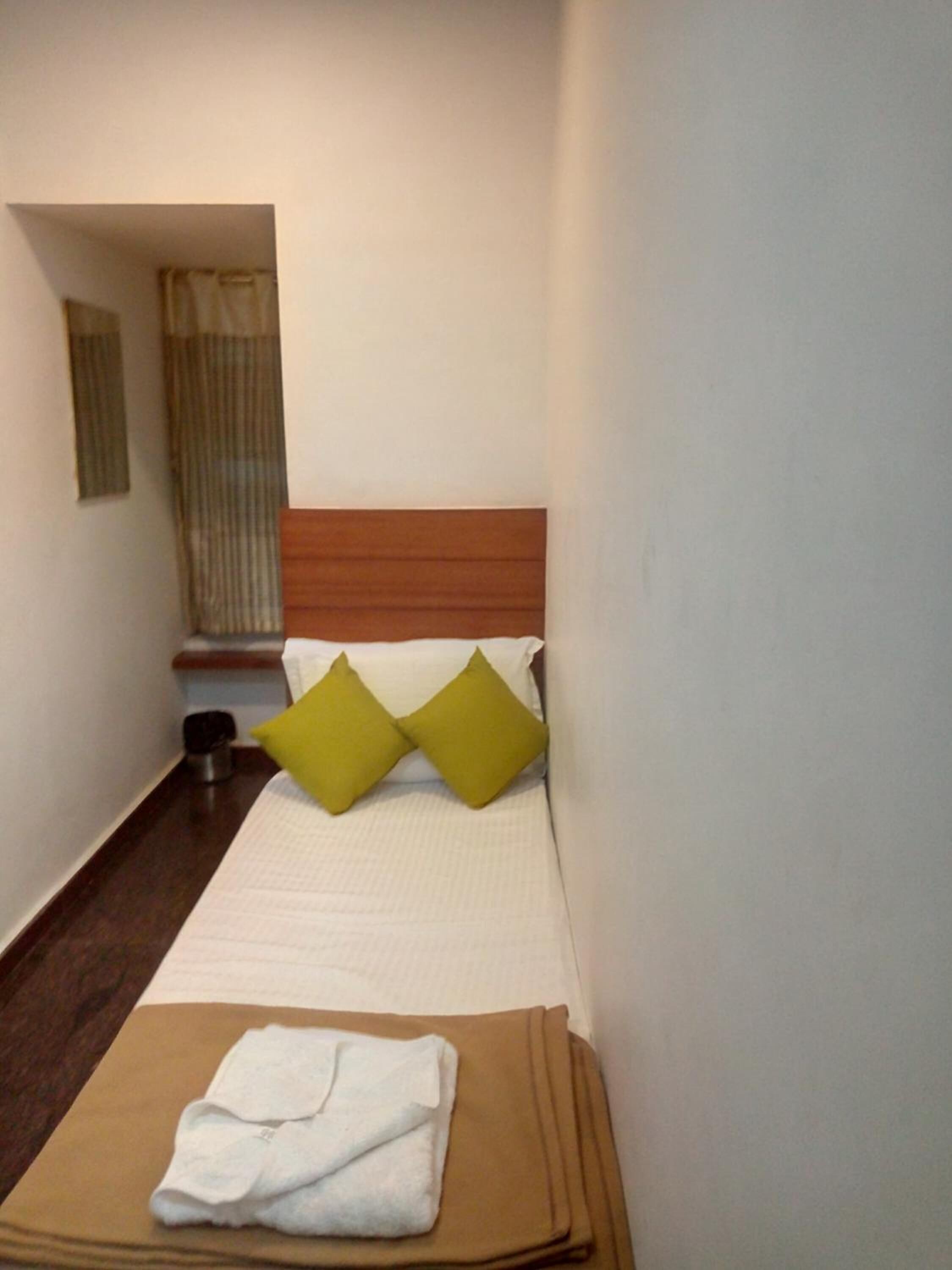 A narrow hotel room with a bed, two pillows, a folded towel, and a small table with a cup.