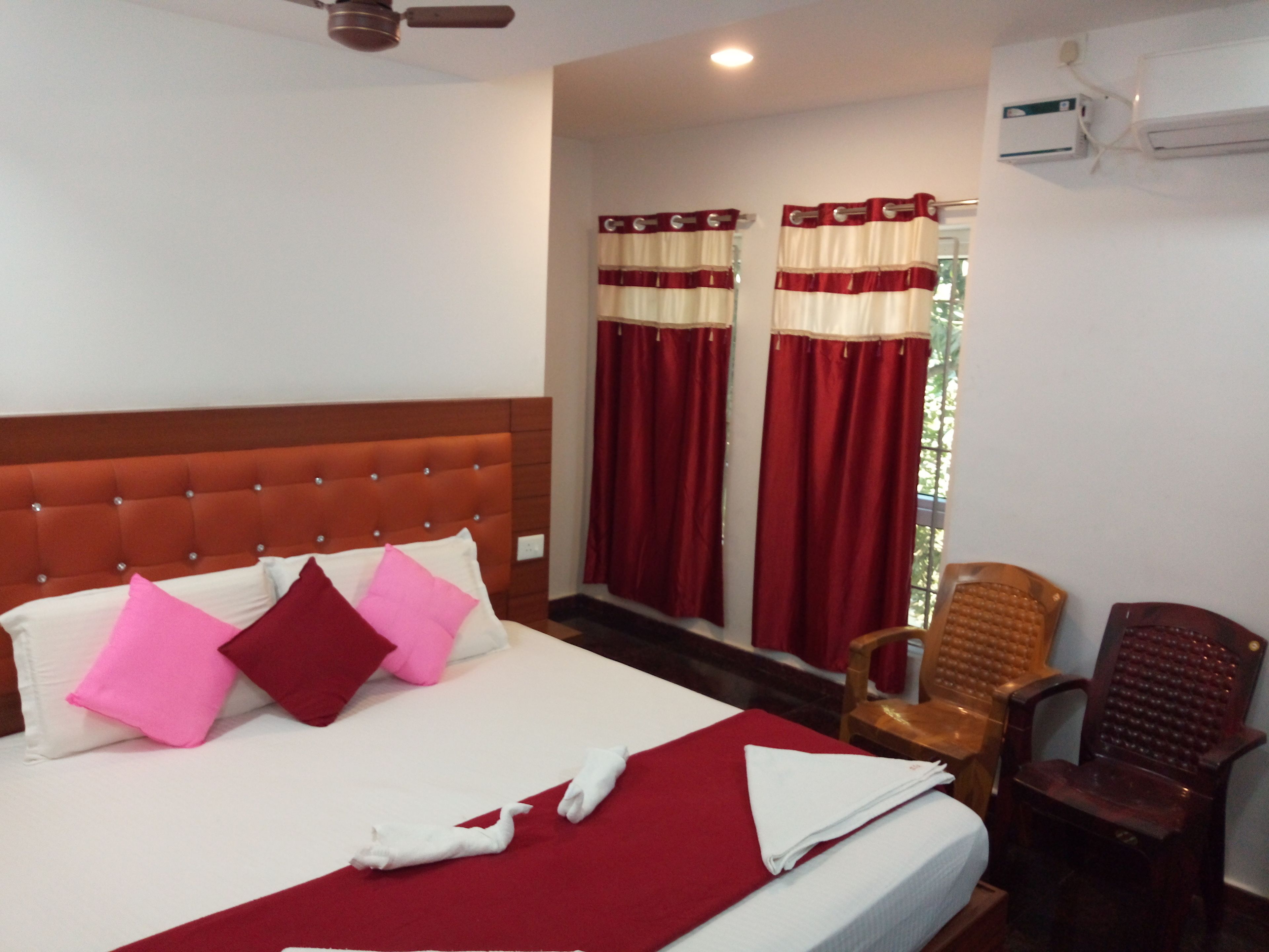 A hotel room with a large bed, red and white curtains, a wooden headboard, a wooden chair, and an air conditioning unit.