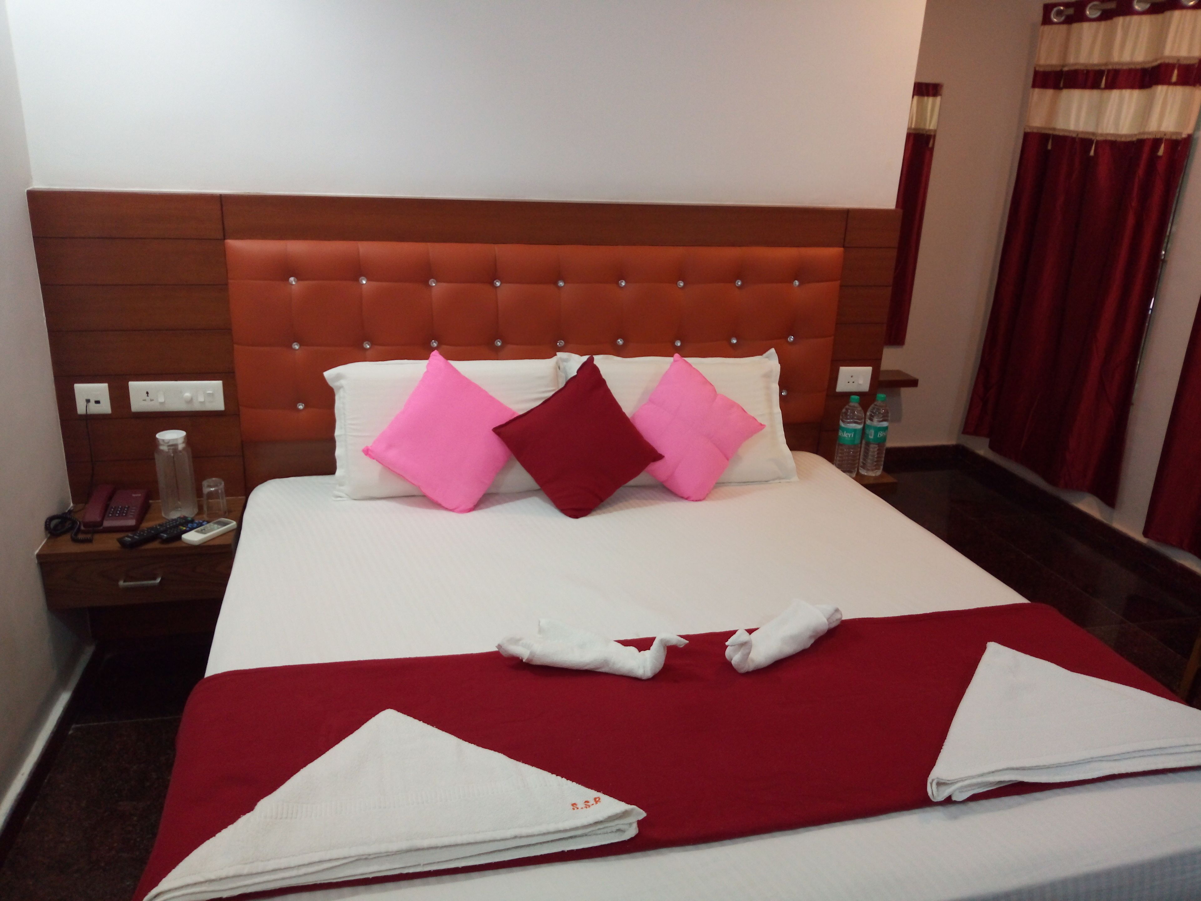 Una habitación de hotel con una cama grande, ropa de cama roja y blanca, un cabecero de madera y una mesita de noche con un teléfono, controles remotos y una botella de agua.