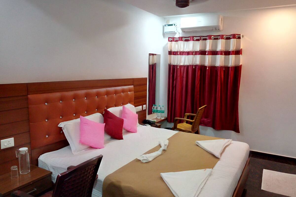 A hotel room with a large bed, a wooden headboard, a desk with a chair, a small table with bottles, and a window with red curtains.