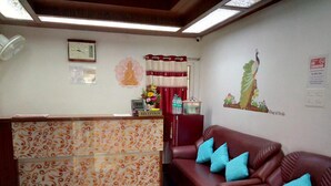 Un área de recepción con un mostrador decorado, un reloj, una estatua de Buda montada en la pared, una cortina roja, un sofá rojo con cojines azules y un letrero de 'No Fumar'.