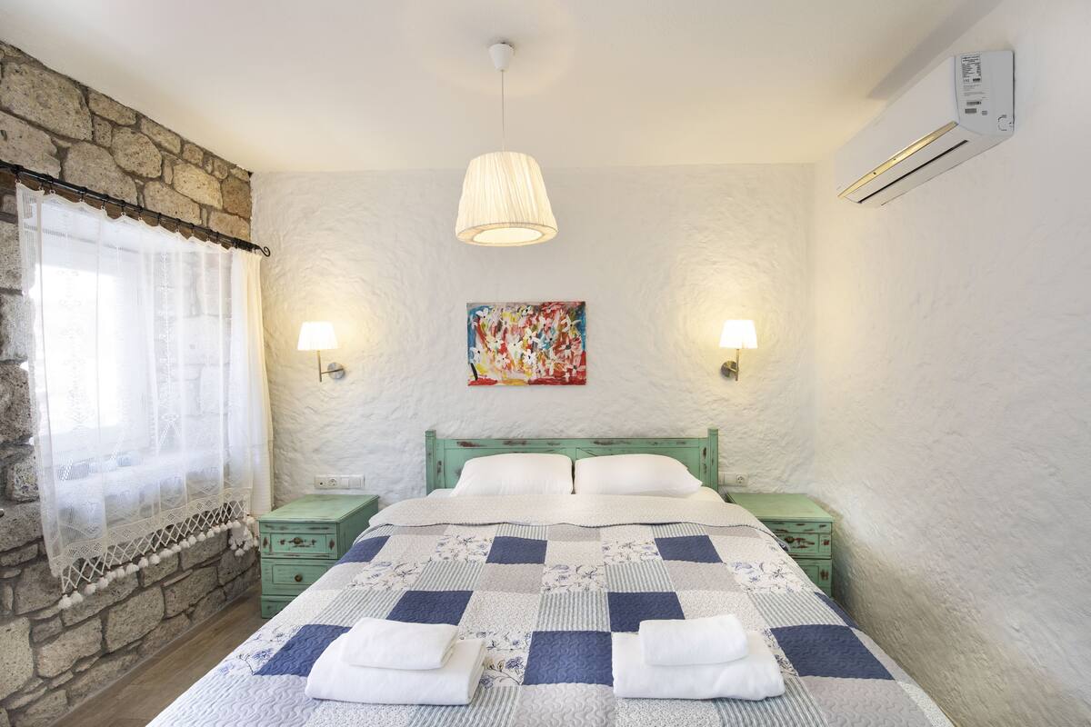Un dormitorio con colcha a cuadros, dos mesitas de noche, una lámpara, un cuadro y una pared de piedra.