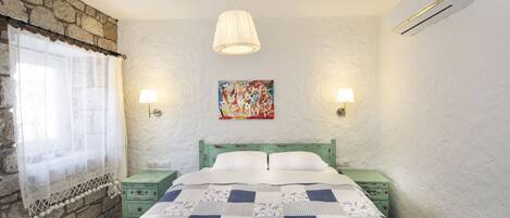 A bedroom with a checkered bedspread, two bedside tables, a nightstand, a lamp, a painting, and a stone wall.