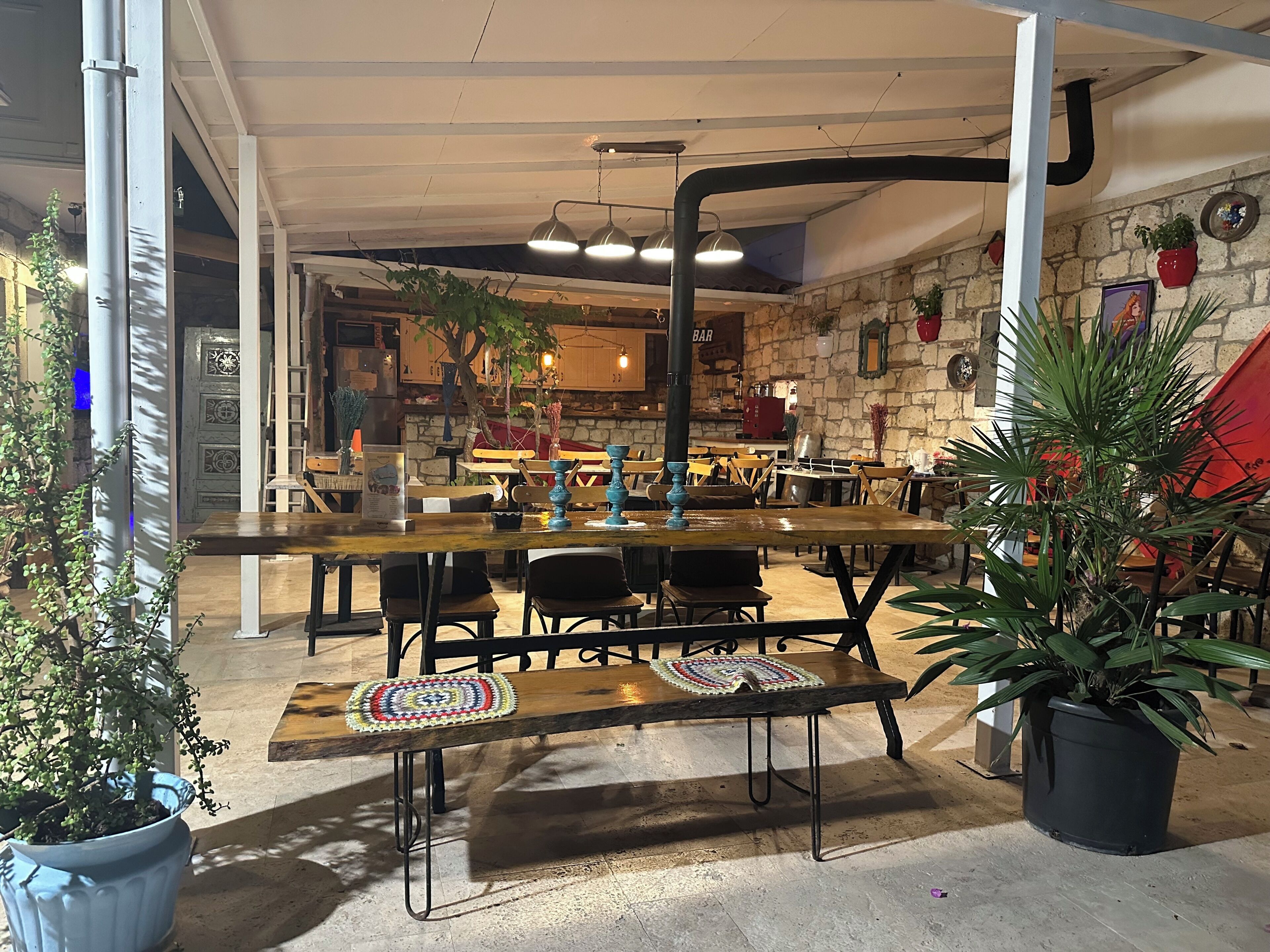 An indoor dining area with wooden tables and chairs, potted plants, and a stone wall.