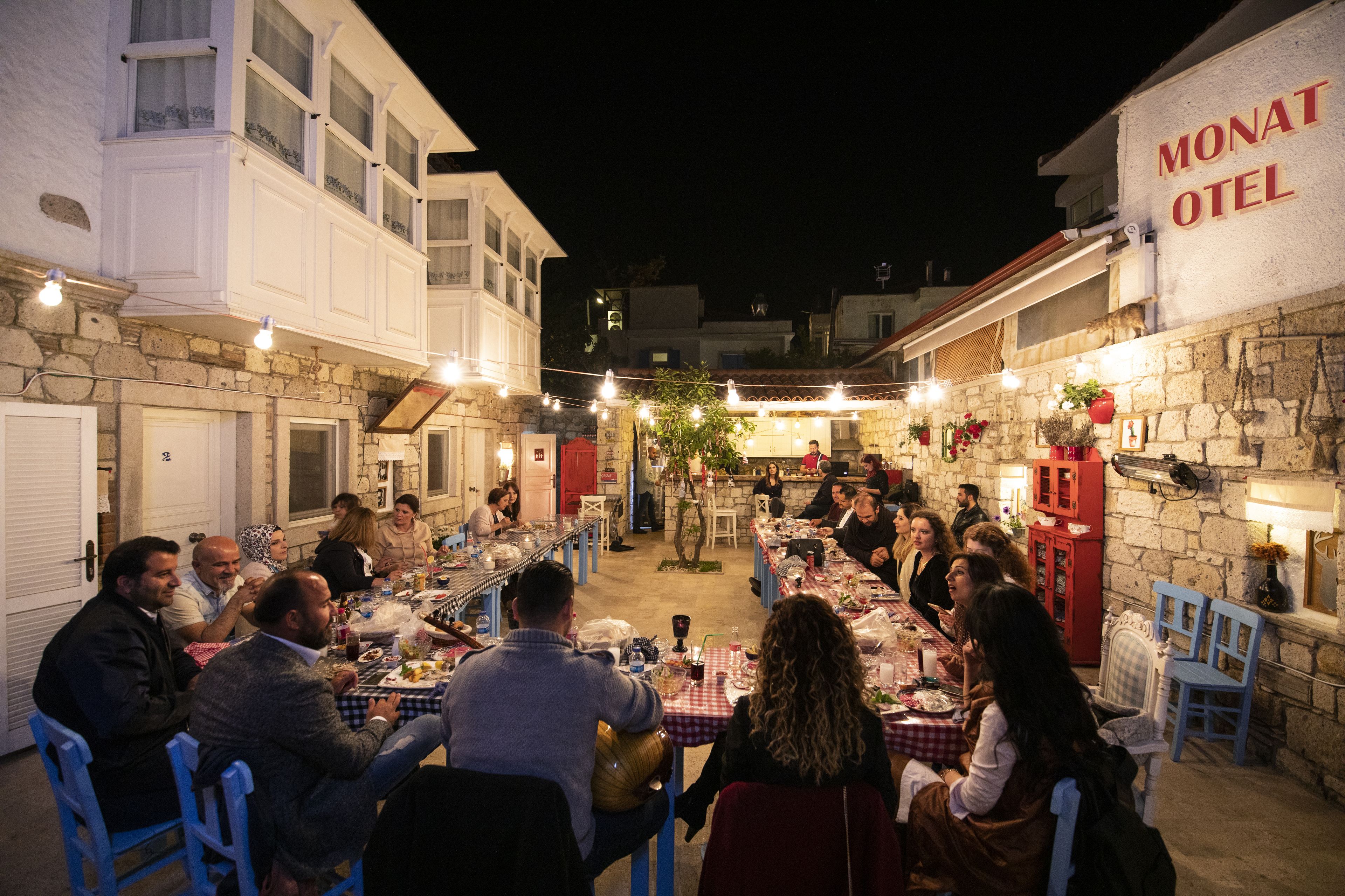 Un área de comedor al aire libre con paredes de piedra y un letrero que dice 'Monat Hotel'. Las mesas están dispuestas con comensales y se ven luces colgantes.