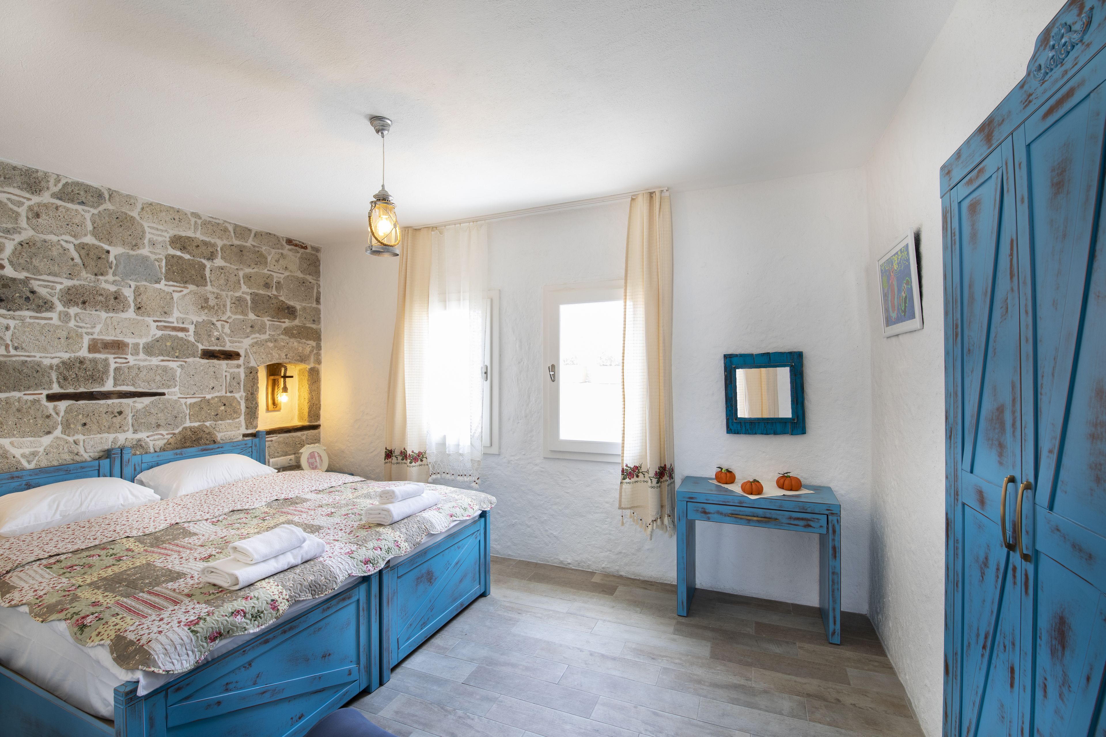 A bedroom with a stone wall, a bed with a floral bedspread, a small table with pumpkins, and a blue wardrobe.