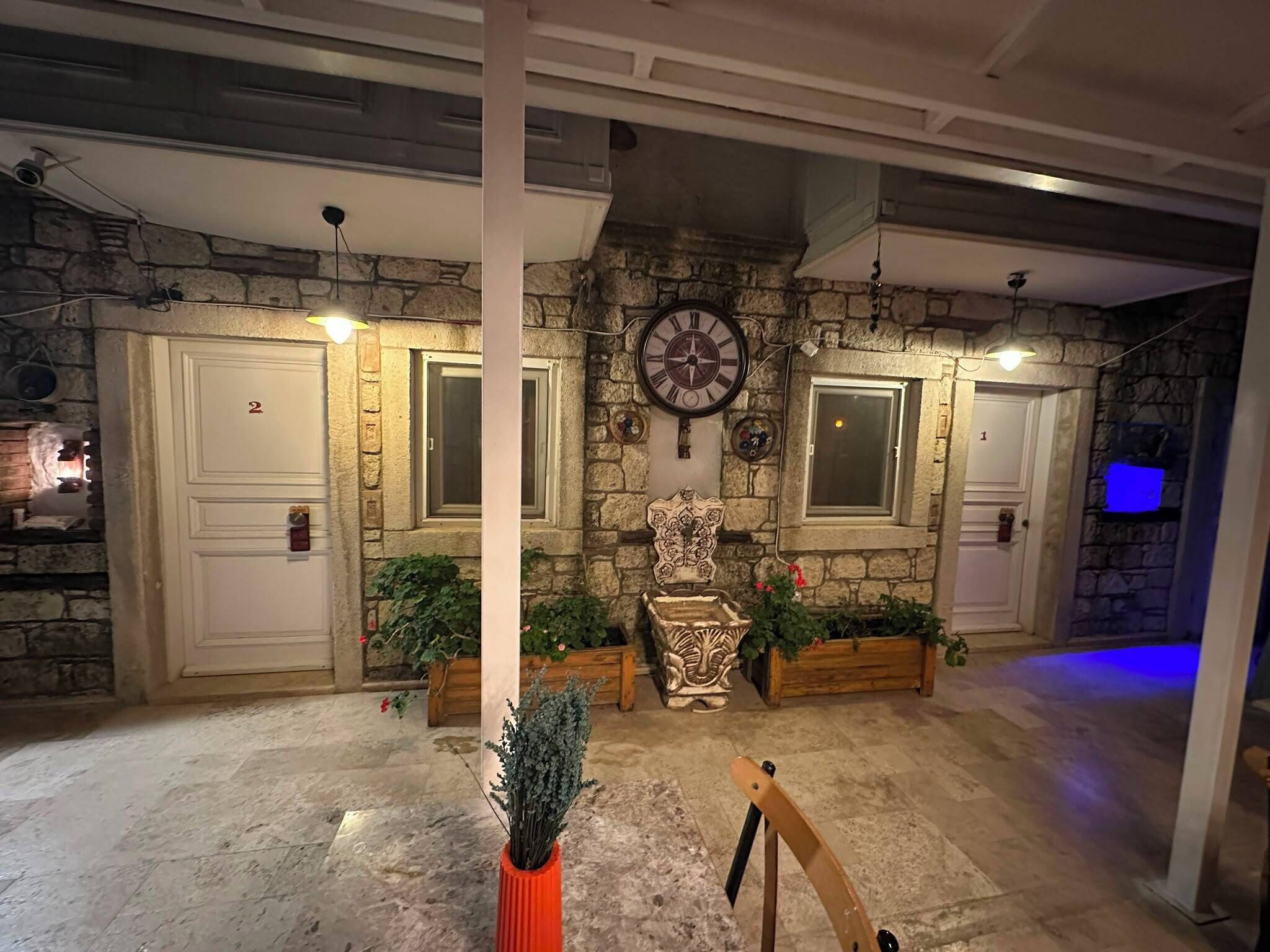 Une pièce aux murs de pierre, avec deux portes blanches, une grande horloge murale et des plantes en pot.