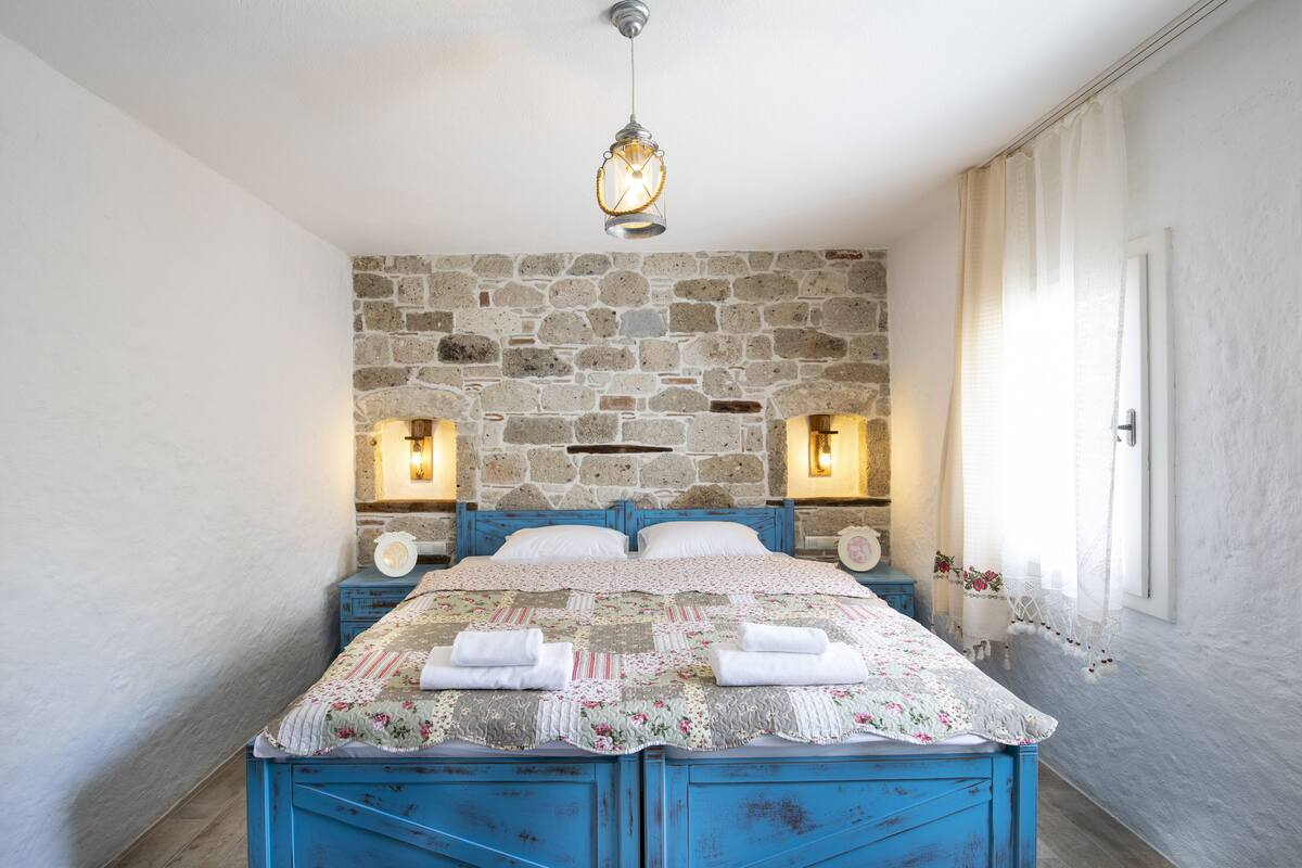 Un dormitorio con pared de piedra, una cama con colcha estampada y dos luces fijadas a la pared.