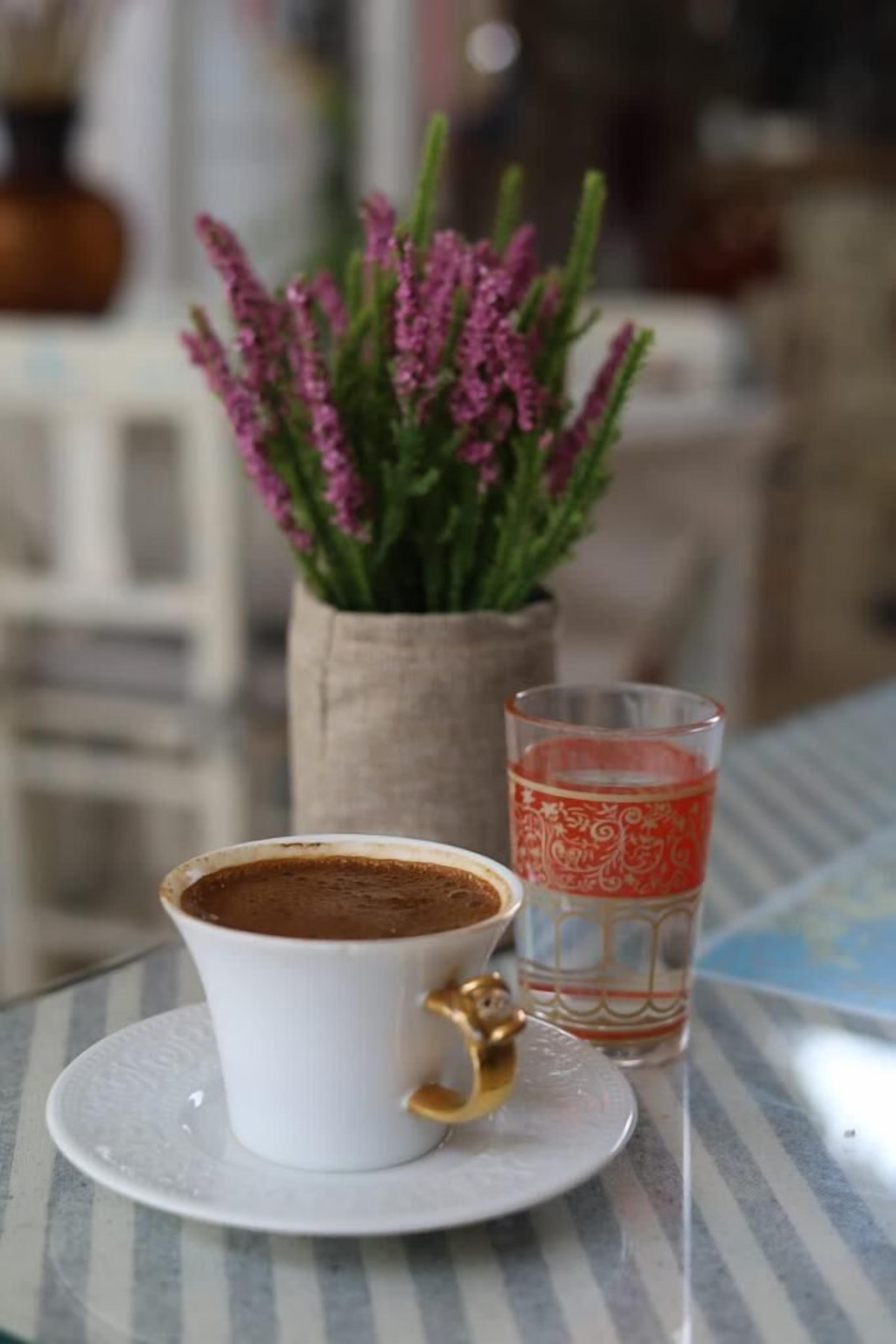 Una taza de café sobre un platito, un vaso de agua y una maceta con flores rosadas sobre una mesa.