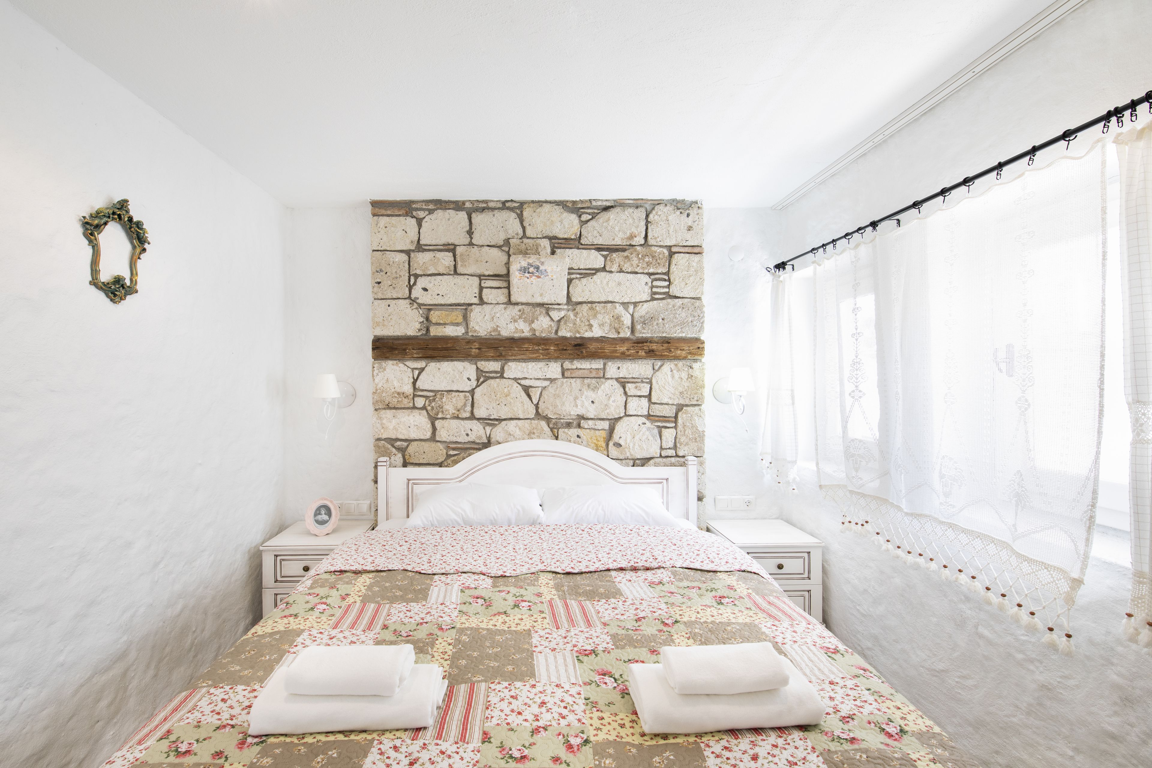 A bedroom with a stone wall, a wooden headboard, a floral bedspread, and a white nightstand with a lamp.