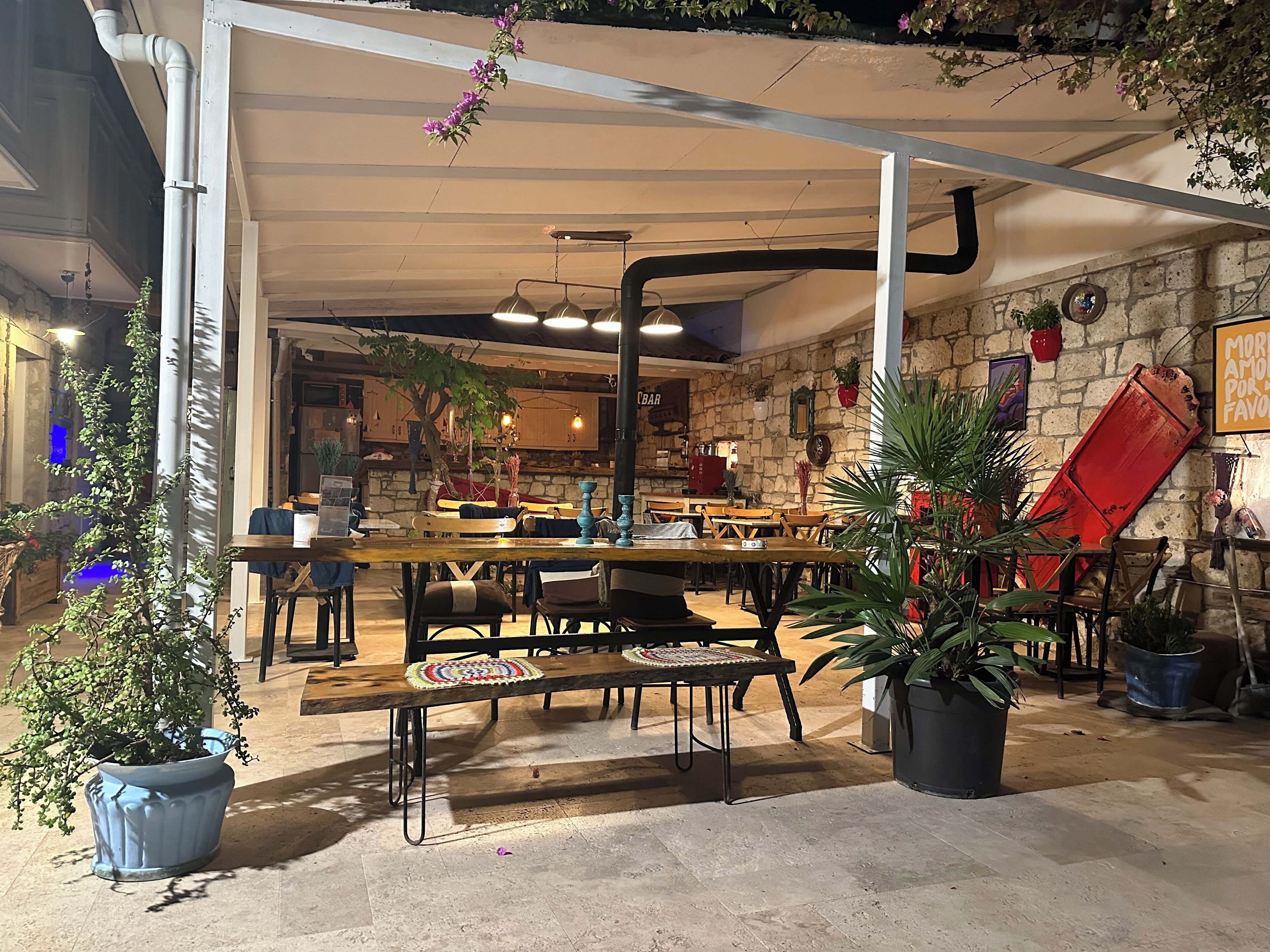 Un café al aire libre con mesas y sillas, rodeado de plantas en macetas y un muro de piedra.