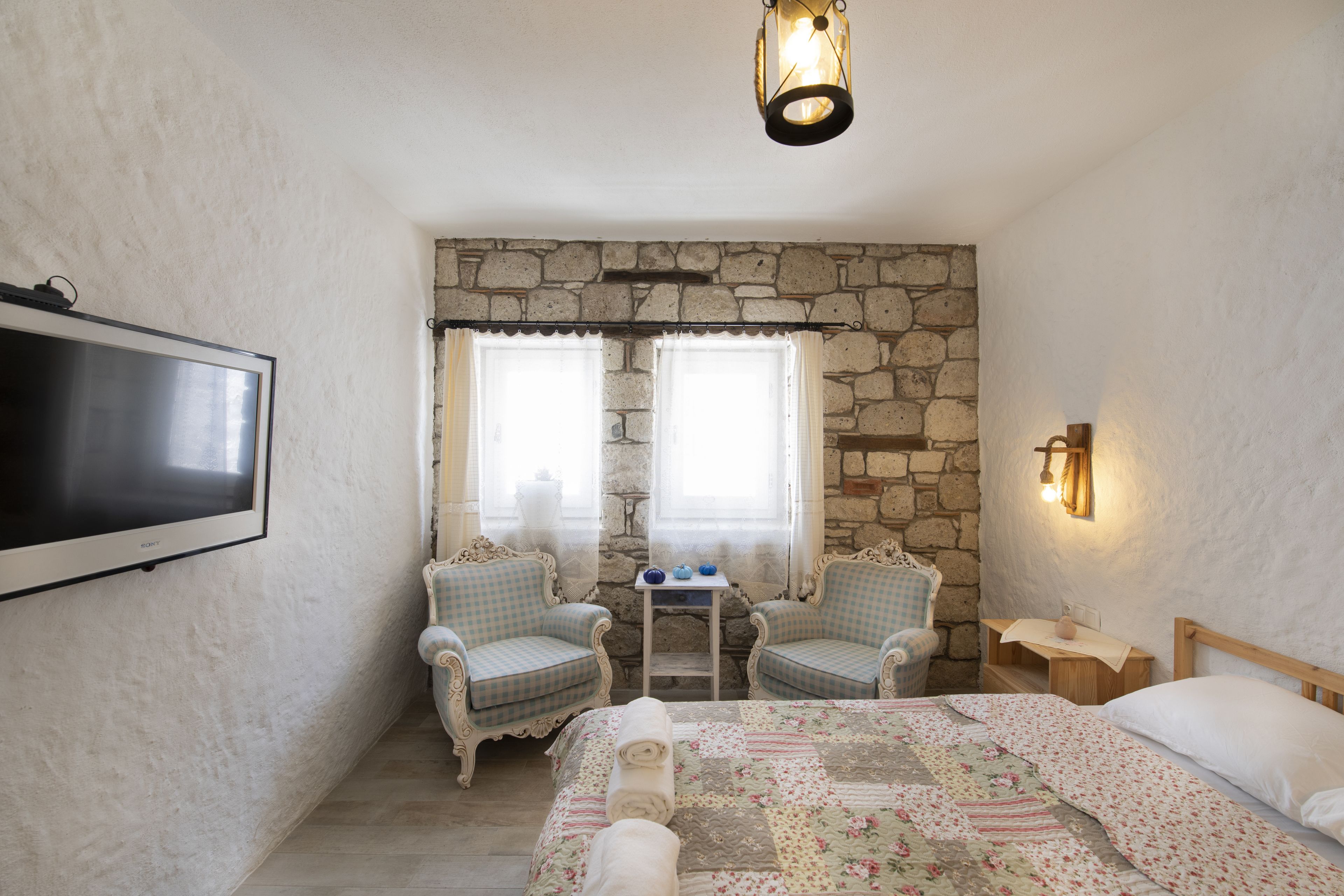 A bedroom with a stone wall, a bed with floral bedding, two checkered armchairs, a small table with blue vases, and a mounted TV.