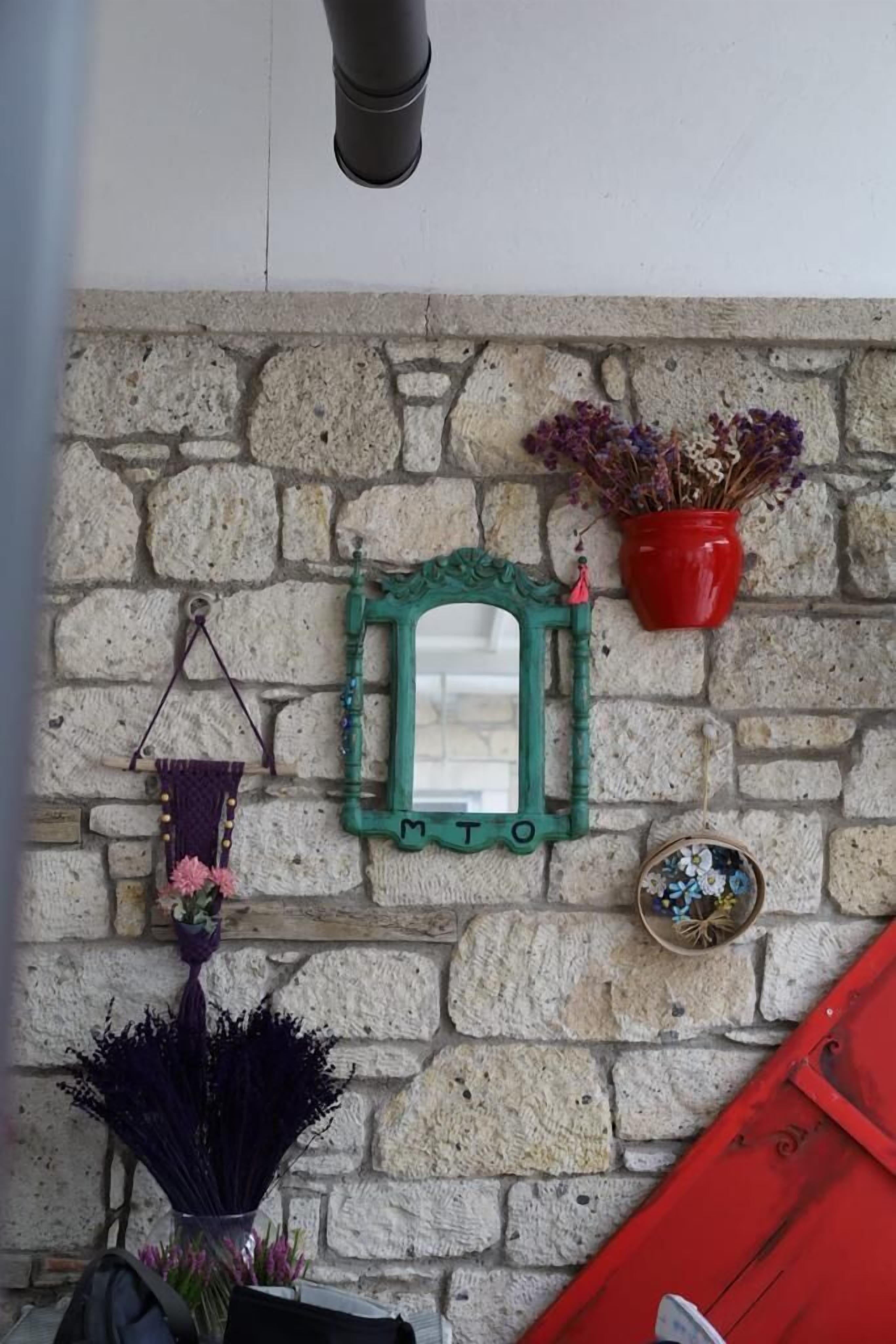 A stone wall with a green-framed mirror, a red vase with dried flowers, and a small decorative bowl.