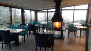 Ein Restaurant mit einem zentralen Kamin, Tischen und Stühlen zum Essen und großen Fenstern mit Blick ins Freie.