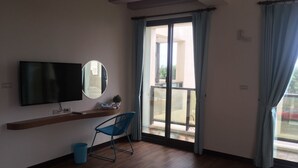 Una habitación con un televisor de pantalla plana montado en la pared, una silla azul, una mesita con una planta y un ventanal corredero que da a un balcón.