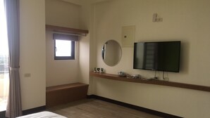 Habitación de hotel con cama, televisor, ventana con cortinas, espejo y un pequeño estante con objetos.