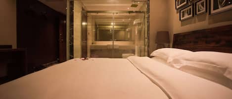 A hotel room with a large bed, a glass-enclosed shower, a wooden headboard, and framed pictures on the wall.