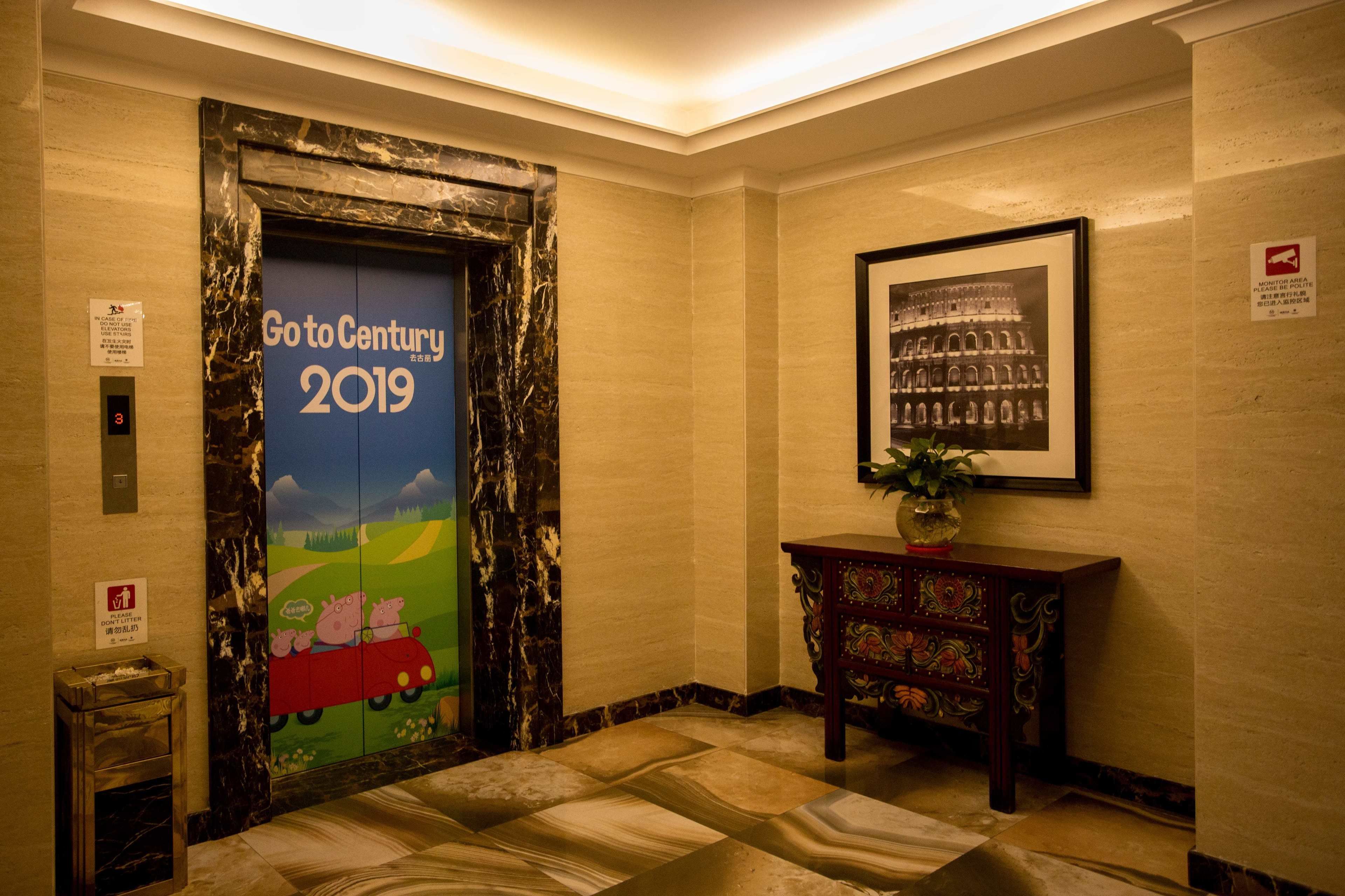 A hotel lobby with a marble floor, a door with a 'Go to Century 2019' mural, a framed picture on the wall, and a small trash bin.