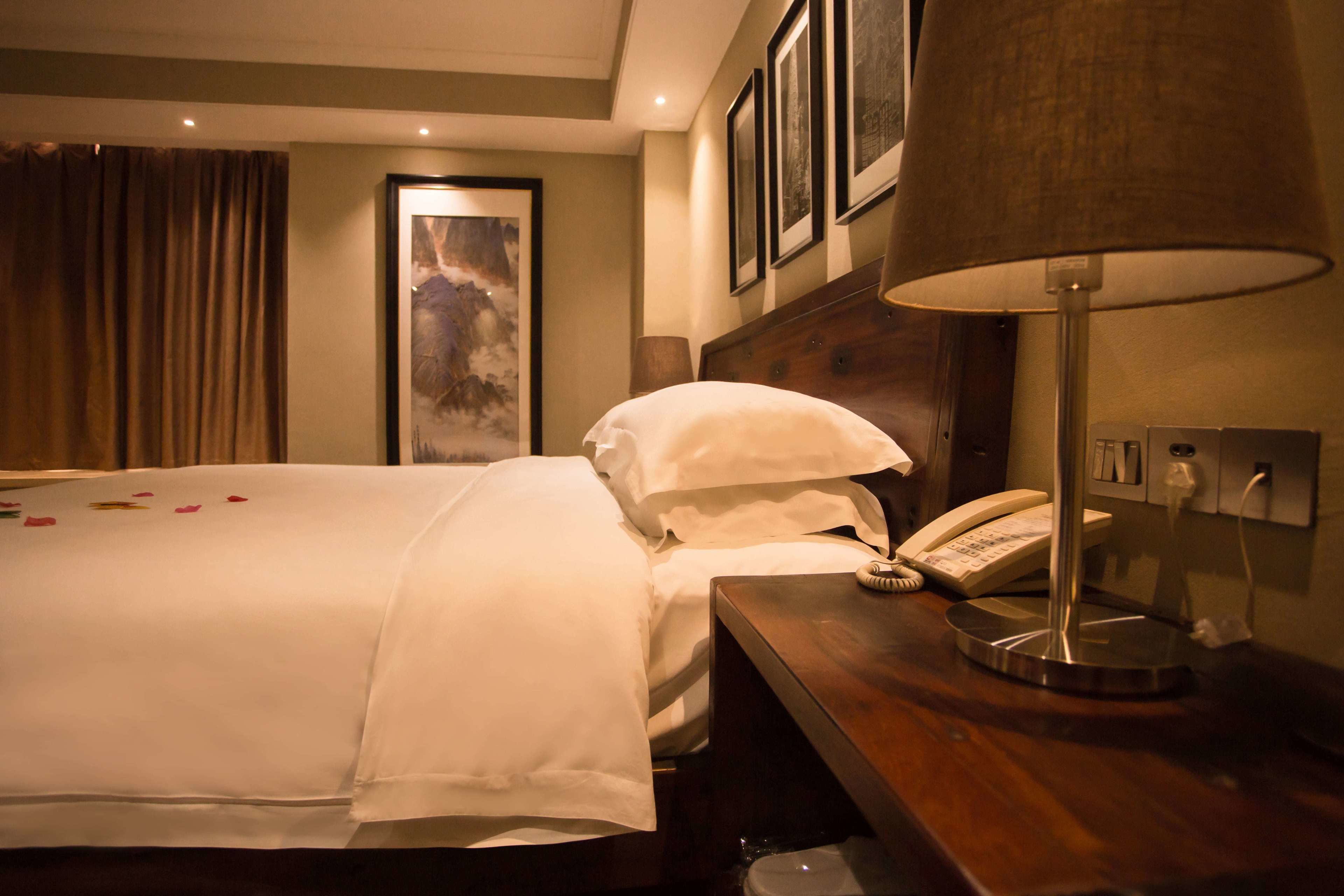 A hotel room with a neatly made bed, a bedside telephone, and a lamp on a wooden nightstand.