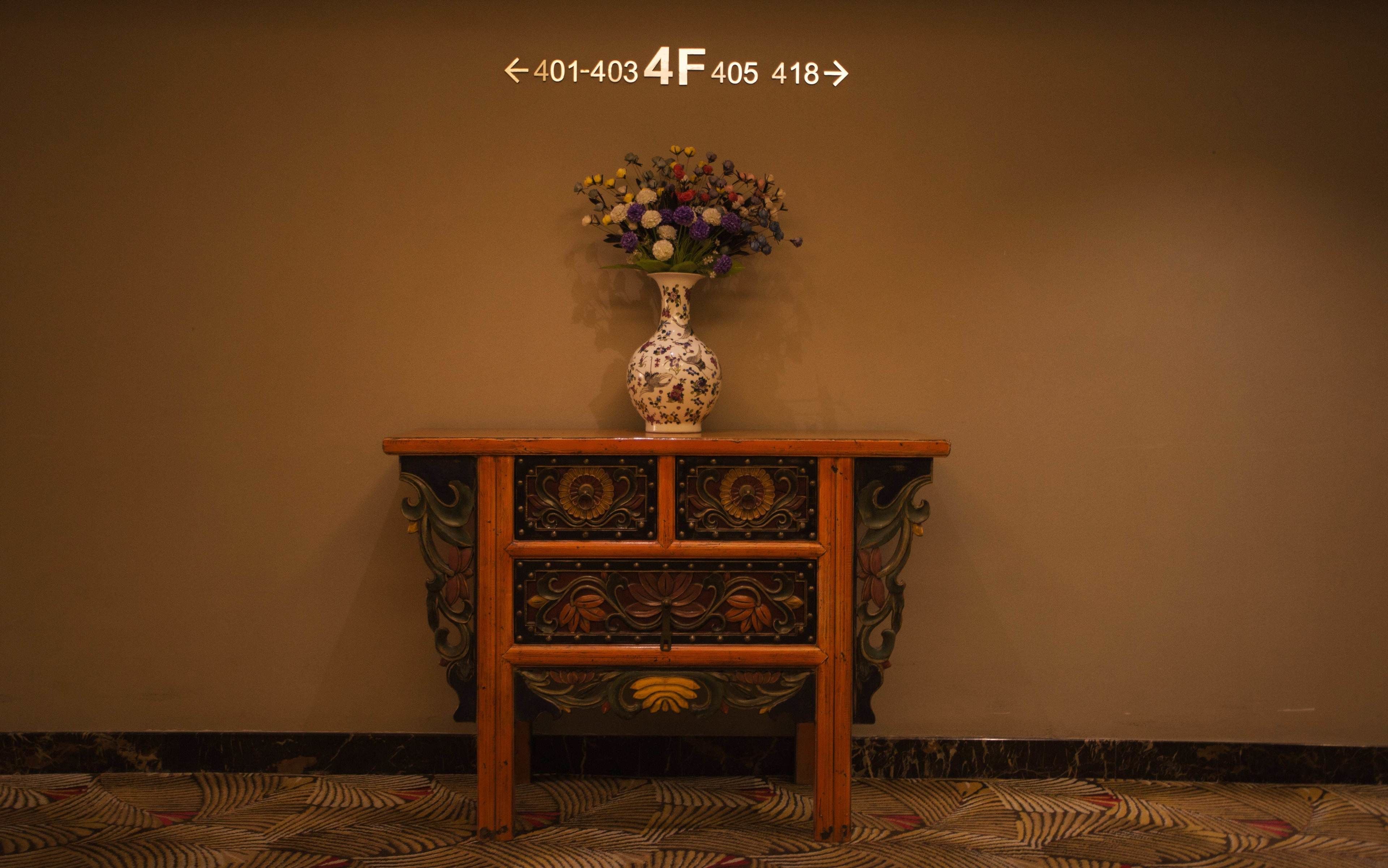 Um console de madeira com um vaso de flores em cima, posicionado em frente a uma parede lisa com um tapete estampado.